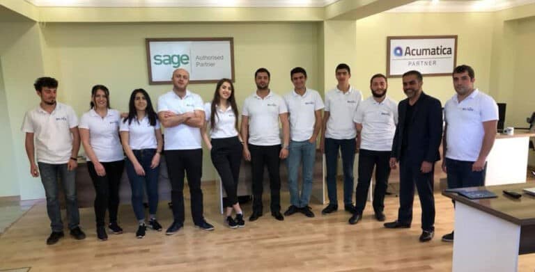 Group of staff in white polo shirts pose in an office with Sage Authorized Partner and Acumatica Partner signs on the wall