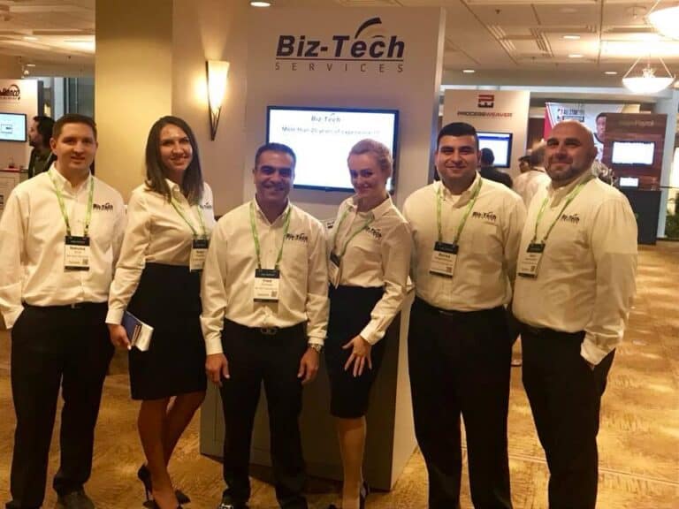 Six Biz-Tech Services team members in branded white shirts stand at a trade show booth with Biz-Tech Services signage and display screens behind them