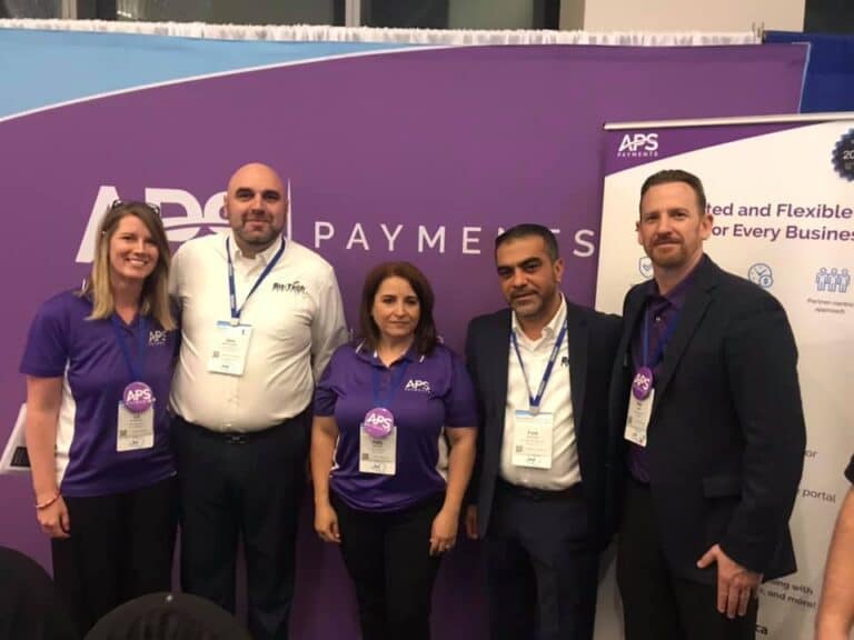 Five people wearing conference badges stand in front of an APS Payments branded booth backdrop